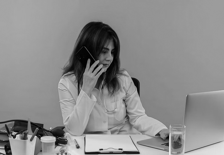A female doctor in a white coat talks on the phone with the Emergency Help Desk while working on a laptop at a desk with medical tools, papers, and a glass of water, set against an orange background.