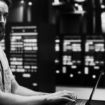 A man with a beard, wearing a light shirt and lanyard, works on a laptop in a server room filled with illuminated servers—demonstrating how Server solutions Portland can enhance productivity.