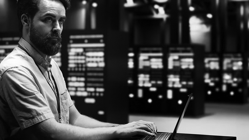 A man with a beard, wearing a light shirt and lanyard, works on a laptop in a server room filled with illuminated servers—demonstrating how Server solutions Portland can enhance productivity.