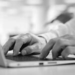 Close-up of a person’s hands typing on a laptop keyboard, with a blurred background including greenery and light, suggesting a bright indoor workspace—ideal for business tasks or using a cloud-based collaboration tool.