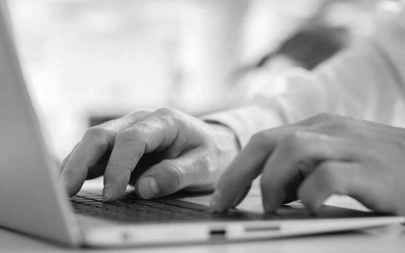 Close-up of a person’s hands typing on a laptop keyboard, with a blurred background including greenery and light, suggesting a bright indoor workspace—ideal for business tasks or using a cloud-based collaboration tool.