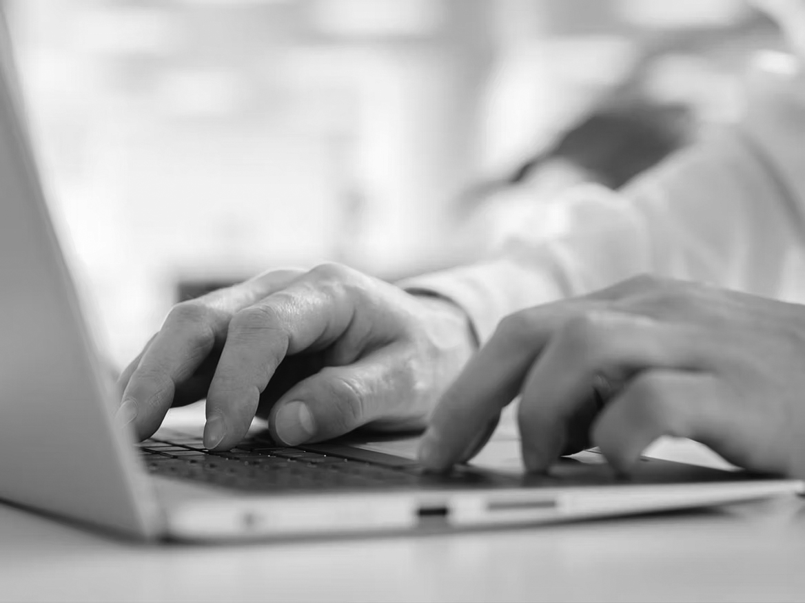 Close-up of a person’s hands typing on a laptop keyboard, with a blurred background including greenery and light, suggesting a bright indoor workspace—ideal for business tasks or using a cloud-based collaboration tool.