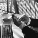 A person in a suit uses a smartphone while sitting at a table with an open laptop and a cup of coffee, sunlight streaming through window blinds—highlighting the importance of privacy and ethics in today’s connected workspaces.