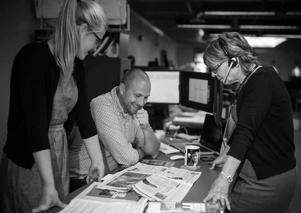 Three people in an office discuss printed newspapers and papers spread on a desk, exploring deployment solutions; two women stand on either side, while a man sits in the center, smiling. Computers and office supplies are visible.