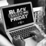 A person uses a laptop displaying a Black Friday Sale graphic with a red arrow, shopping cart icon, and Buy button, highlighting an online shopping event and the importance of safe online shopping for secure holidays.