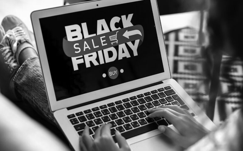 A person uses a laptop displaying a Black Friday Sale graphic with a red arrow, shopping cart icon, and Buy button, highlighting an online shopping event and the importance of safe online shopping for secure holidays.