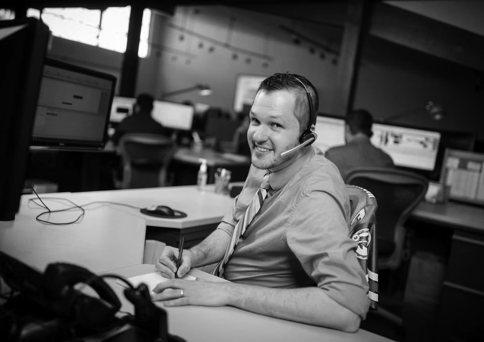 A smiling man wearing a headset and bright blue shirt sits at a desk in a Portland office, writing on paper while looking at the camera. Computer monitors and colleagues working on network solutions are visible in the background.