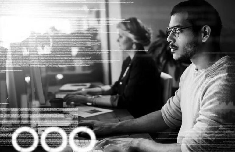 A man and a woman work at computers in an office. The man, focused in glasses and a light sweater, is surrounded by bright digital code—capturing the fast-paced environment of a Managed Services Provider working to minimize IT downtime.