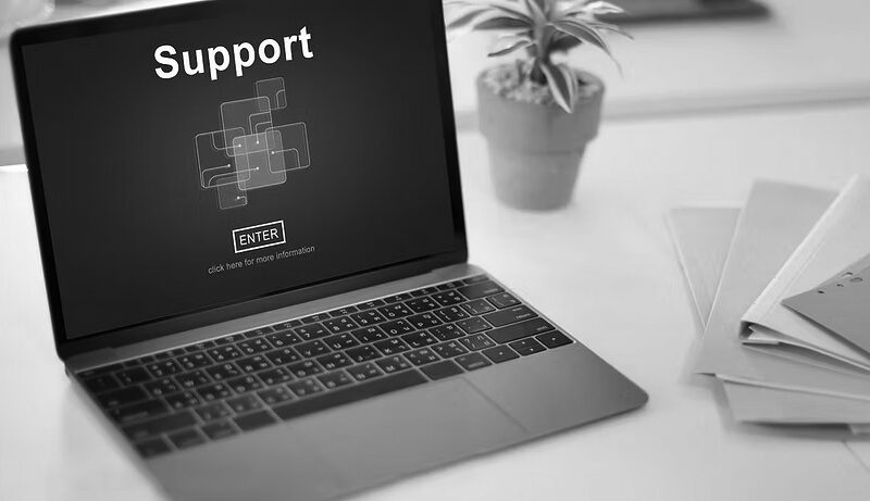 A laptop displays a screen with the word Support and a blue graphic, highlighting Microsoft 365 professional support, sitting on a white desk next to a potted plant and a stack of colored folders.