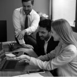 Three colleagues in business attire gather around a wooden desk, discussing IT solutions while reviewing information on a laptop. Office supplies and smartphones are visible on the desk in this modern setting, emphasizing their focus on business potential.