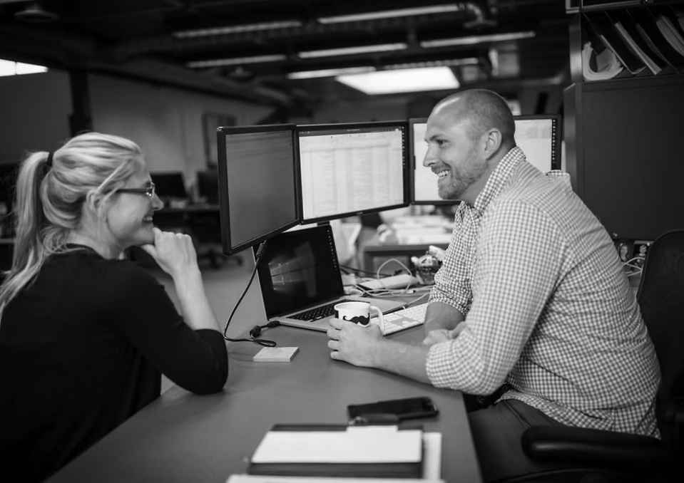 Two coworkers sit at a desk in a modern office, smiling and talking about Vendor Management Services. The man holds a mug while facing a woman, with computer monitors displaying open documents and a notepad on the desk.