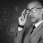A man in a gray suit and glasses stands confidently, touching his glasses, with digital security icons and hexagonal patterns in the background, representing cybersecurity, technology, and Pavelcomm’s expertise in Strategic IT Planning.