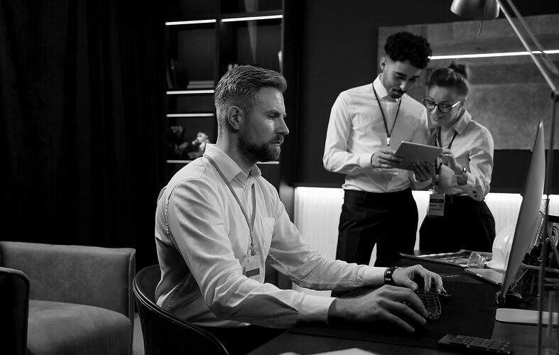 A man with a beard works at a computer in an office, while a man and a woman in business attire discuss something on a tablet in the background, showcasing Managed IT Support for businesses in Portland.