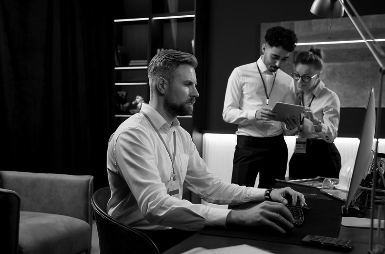 A man with a beard works at a computer in an office, while a man and a woman in business attire discuss something on a tablet in the background, showcasing Managed IT Support for businesses in Portland.