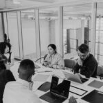 Five people sit around a conference table in a modern office, engaged in discussion about IT solutions to support non-profits. Laptops, notebooks, and papers are on the table, with large windows and glass walls visible in the background.